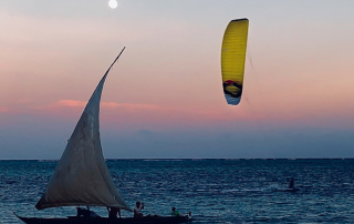 Ein gelber Kite ChronoV3 von Ozonekites am Abendhimmel vor dem Wasser. Im Vordergrund ist ein Segelboot zu sehen. Der Kite ist gut geeignet zum Kitesurfing auf dem Wasser.