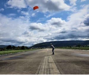 Auf dem Bild sieht man den orangefarbenen Trainerkite IgnitionV3 von Ozonekites. Man kann sich mit dem Kite wie mit einem Segel vom Wind ziehen lassen. Man fährt am auf einem LOngboard.