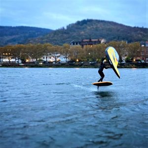 der Kiter fährt auf einem orangefarbenen JKS Board auf dem Fluss Neckar. Der Wind treibt das Wing WASPV2 in der Farbe gelb von Ozonekites an. Deshalb kann der Wingfoiler über das Wasser gleiten.