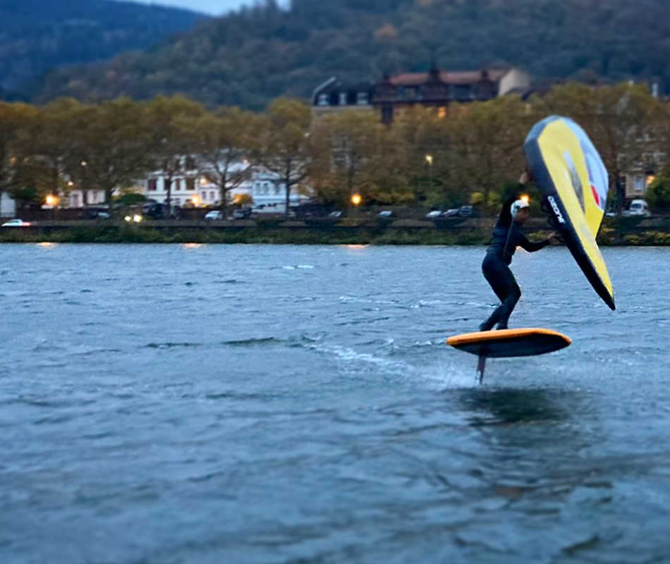 Der Mann fährt auf einem JKS Board auf dem Fluss Neckar. Das Board hat die Farbe orange. Der Wind treibt das Wing WASPV2 in der Farbe gelb von Ozonekites an. Der Wing ist wie ein Segel. Deshalb kann er kiter über das Wasser gleiten